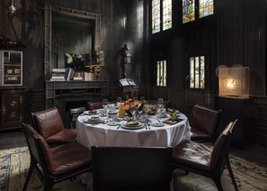 A dining room with a round table set for six, surrounded by leather chairs. There is a bookshelf with books and decorative items, a piano, and a fireplace.
