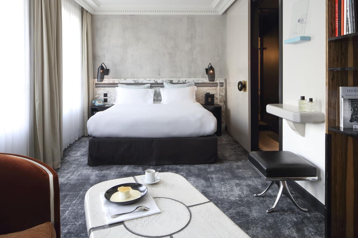 Una habitación de hotel con cama, mesitas de noche, un escritorio con una silla y un pequeño banco.