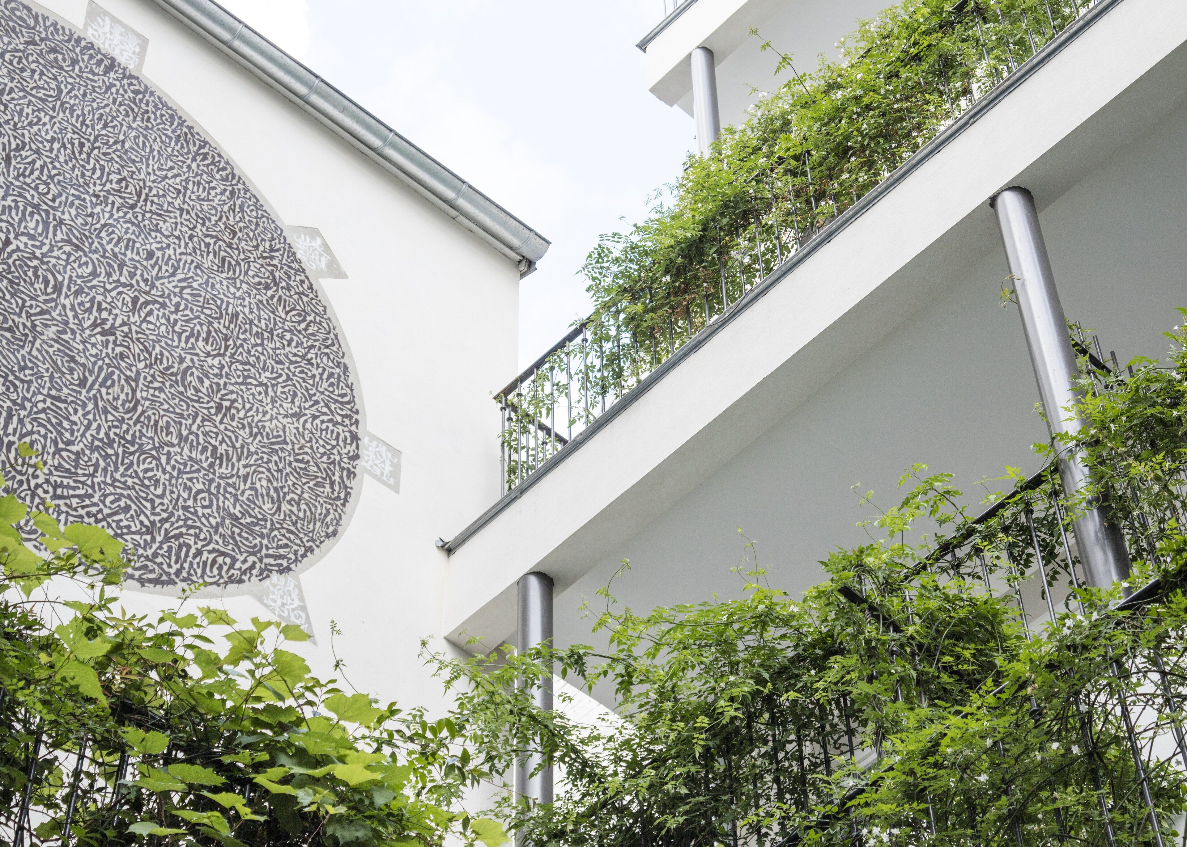 A modern building with a large abstract mural on the left, multiple balconies with greenery, and a clean, white facade.