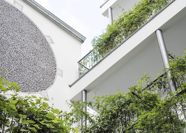 Un edificio moderno con un amplio mural abstracto a la izquierda, varios balcones con vegetación y una fachada blanca y limpia.