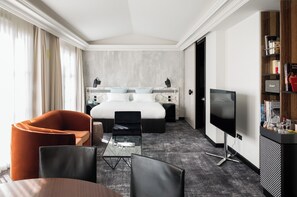 Una habitación de hotel moderna con una cama grande, televisión, zona de estar con mesa y sillas, y una estantería.