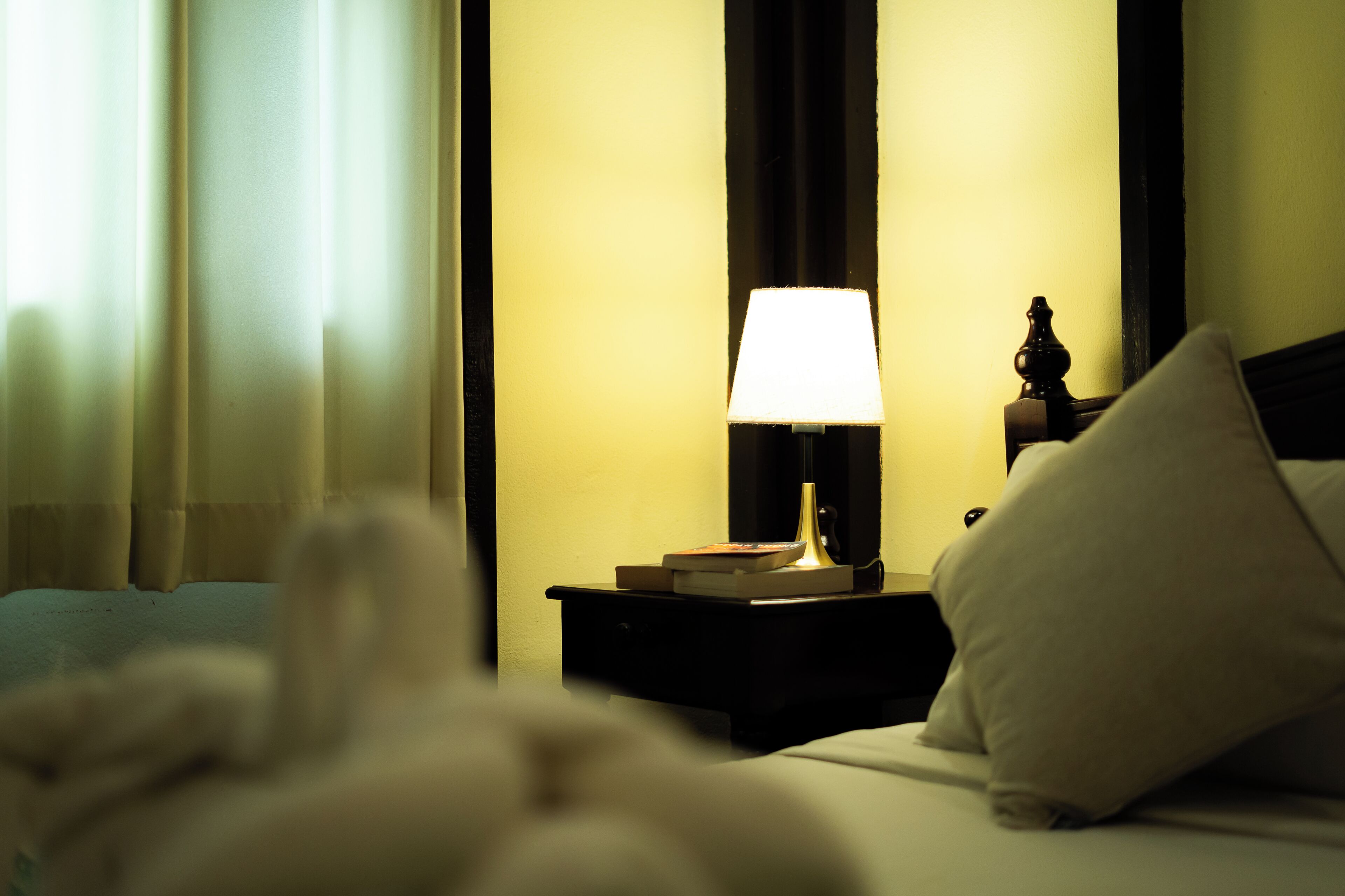 A hotel room with a bed, a bedside table with a lamp, a nightstand, and a pillow.