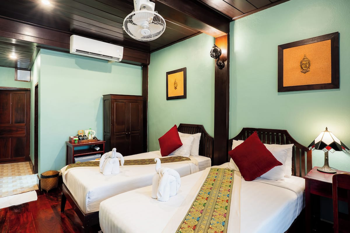 A hotel room with two beds, a wooden headboard, a bedside table with a lamp, an air conditioning unit, and a wall-mounted picture.