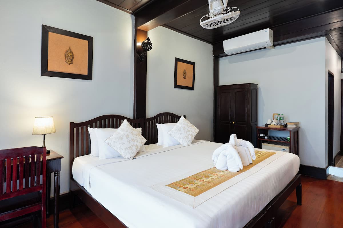 A neatly made bed with white linens, a wooden headboard, and a decorative pillow. There is a small table with a lamp and a red chair to the left.
