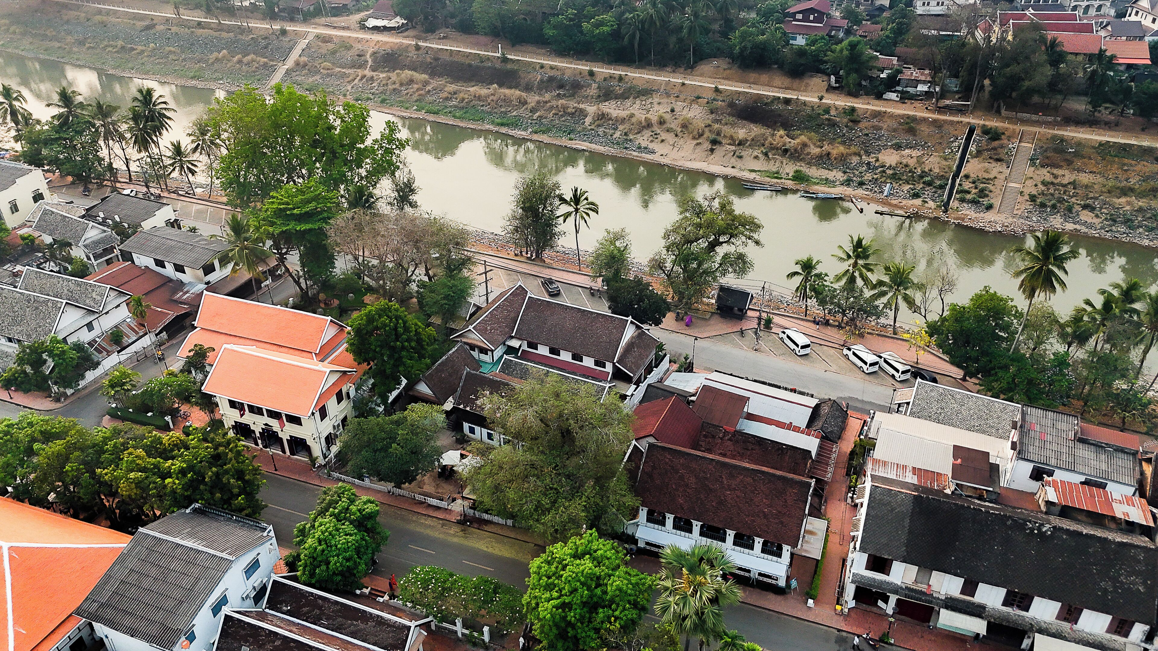 An aerial view of a riverside town with traditional buildings, a paved road, and a bridge.