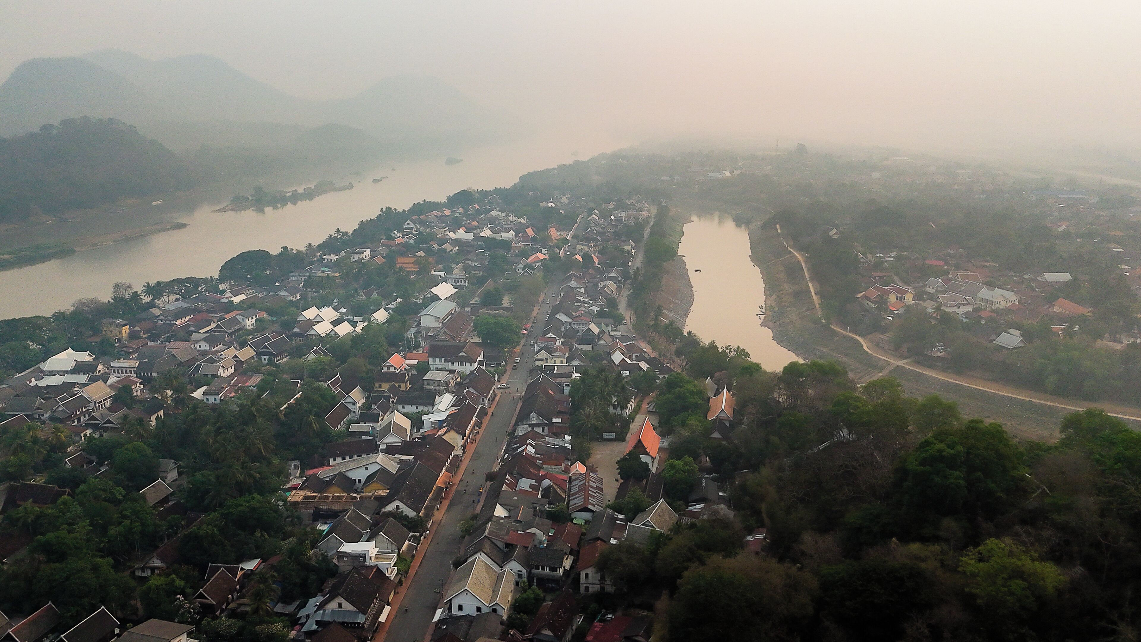 Vista aérea de un pueblo a orillas de un río, con una construcción densa, una carretera principal y el río.