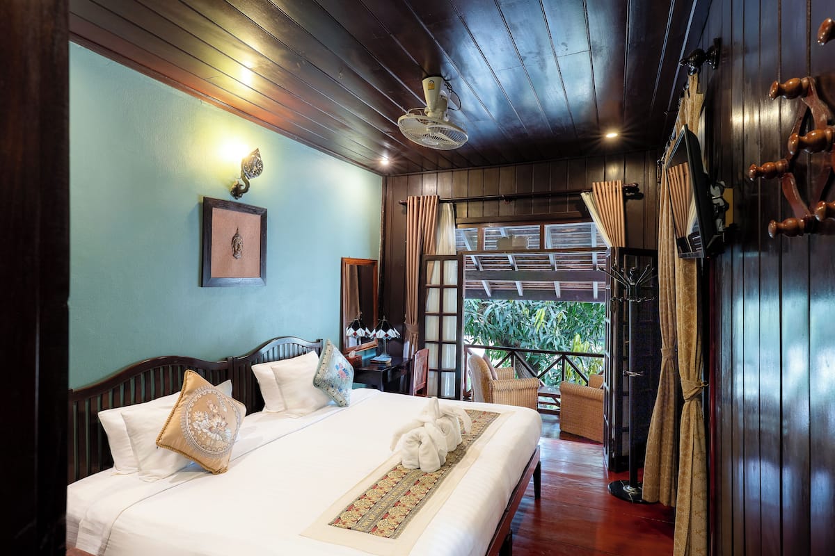 A hotel room with a bed, a desk, a chair, a window with curtains, and a view of greenery outside.