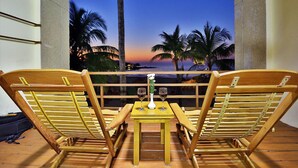 Terraza de madera con dos sillas y una mesa pequeña, con vistas a un atardecer y palmeras.