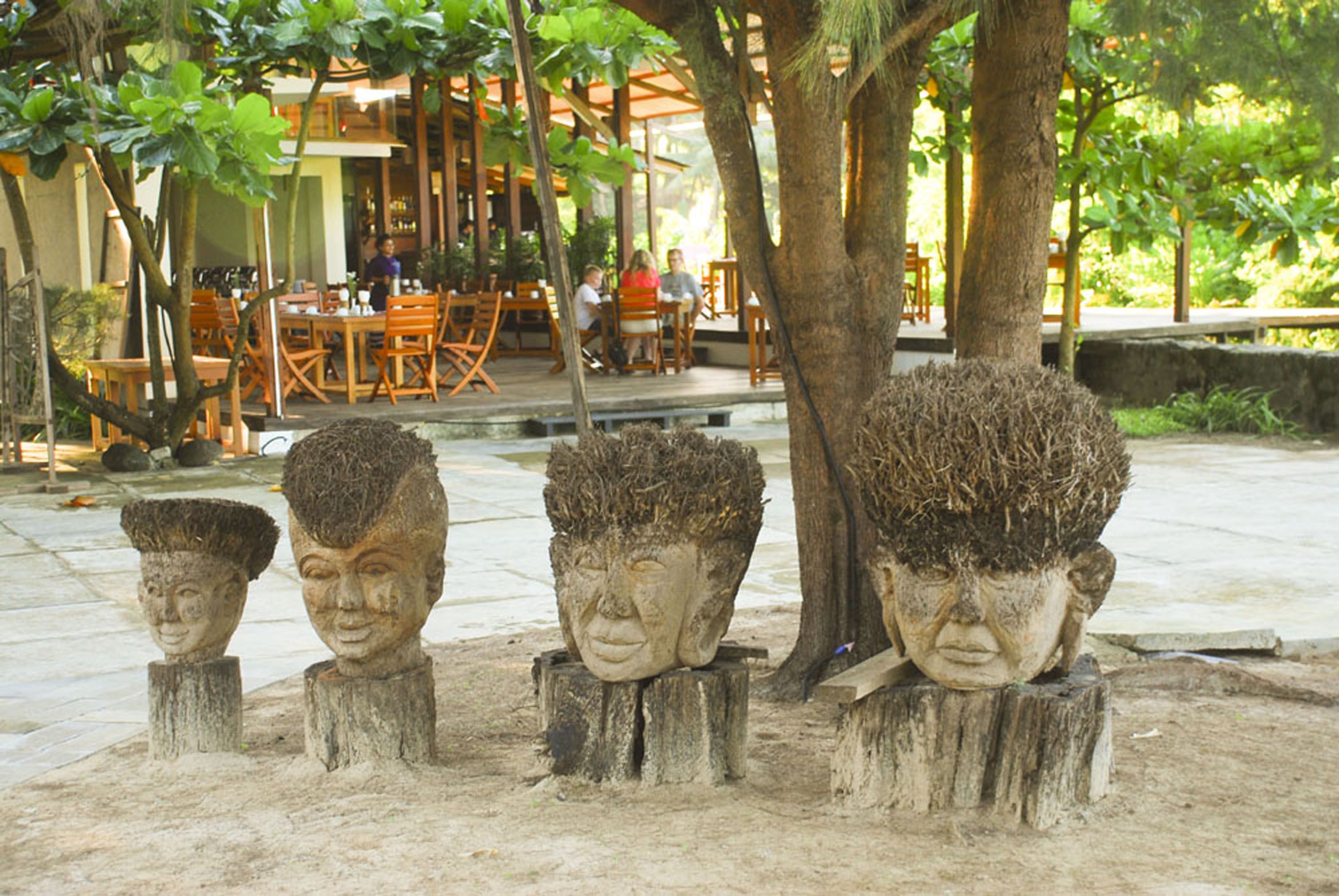 Trois sculptures en pierre, posées sur des souches d’arbres dans un espace extérieur, avec un restaurant en arrière-plan.