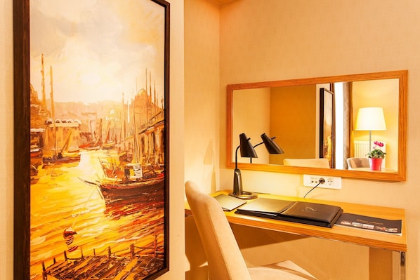 Habitación de hotel con escritorio, silla, lámpara de escritorio, computadora portátil, espejo y una pintura de un paisaje urbano.