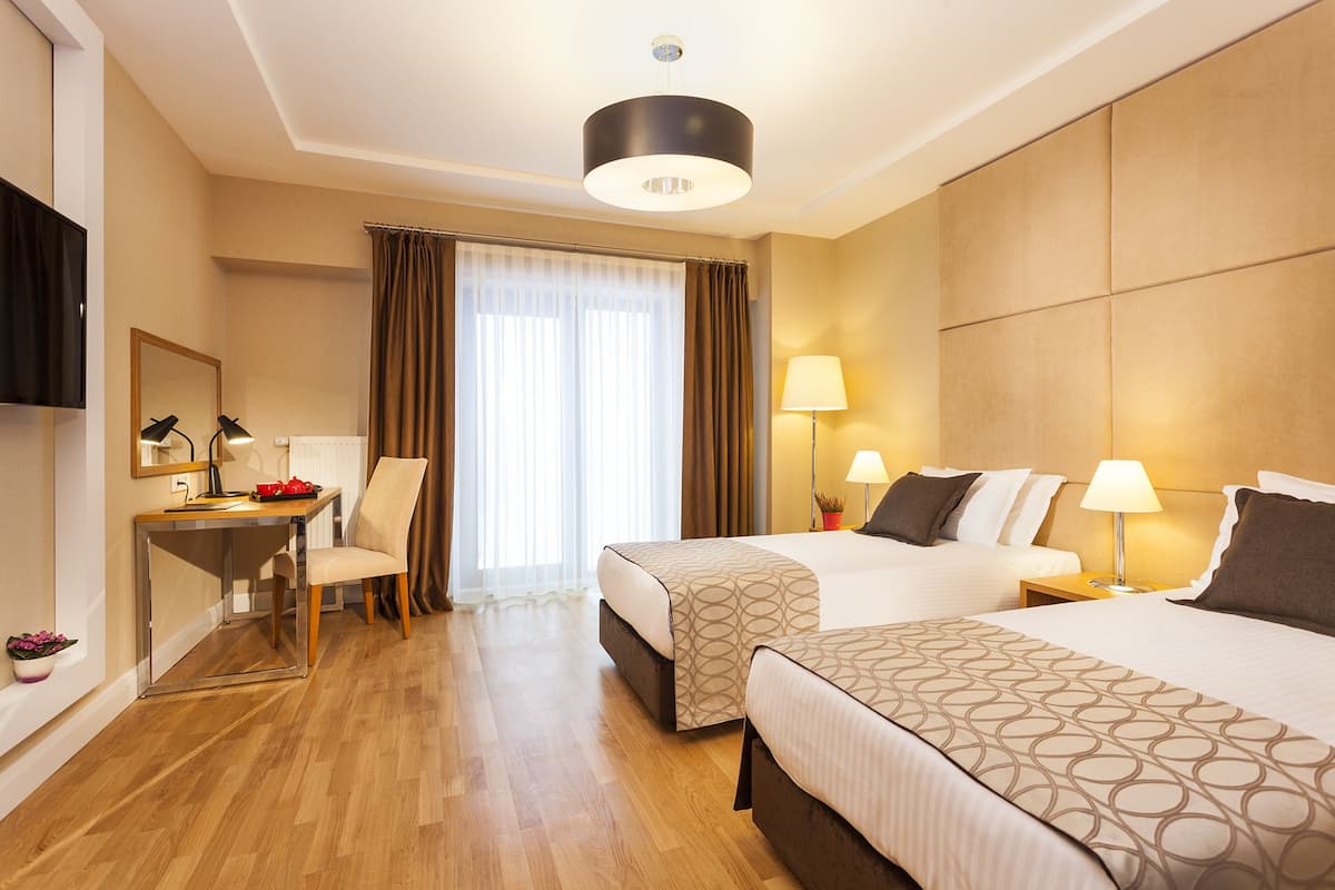 A hotel room with two beds, a desk, a chair, a TV, and a large window with curtains.