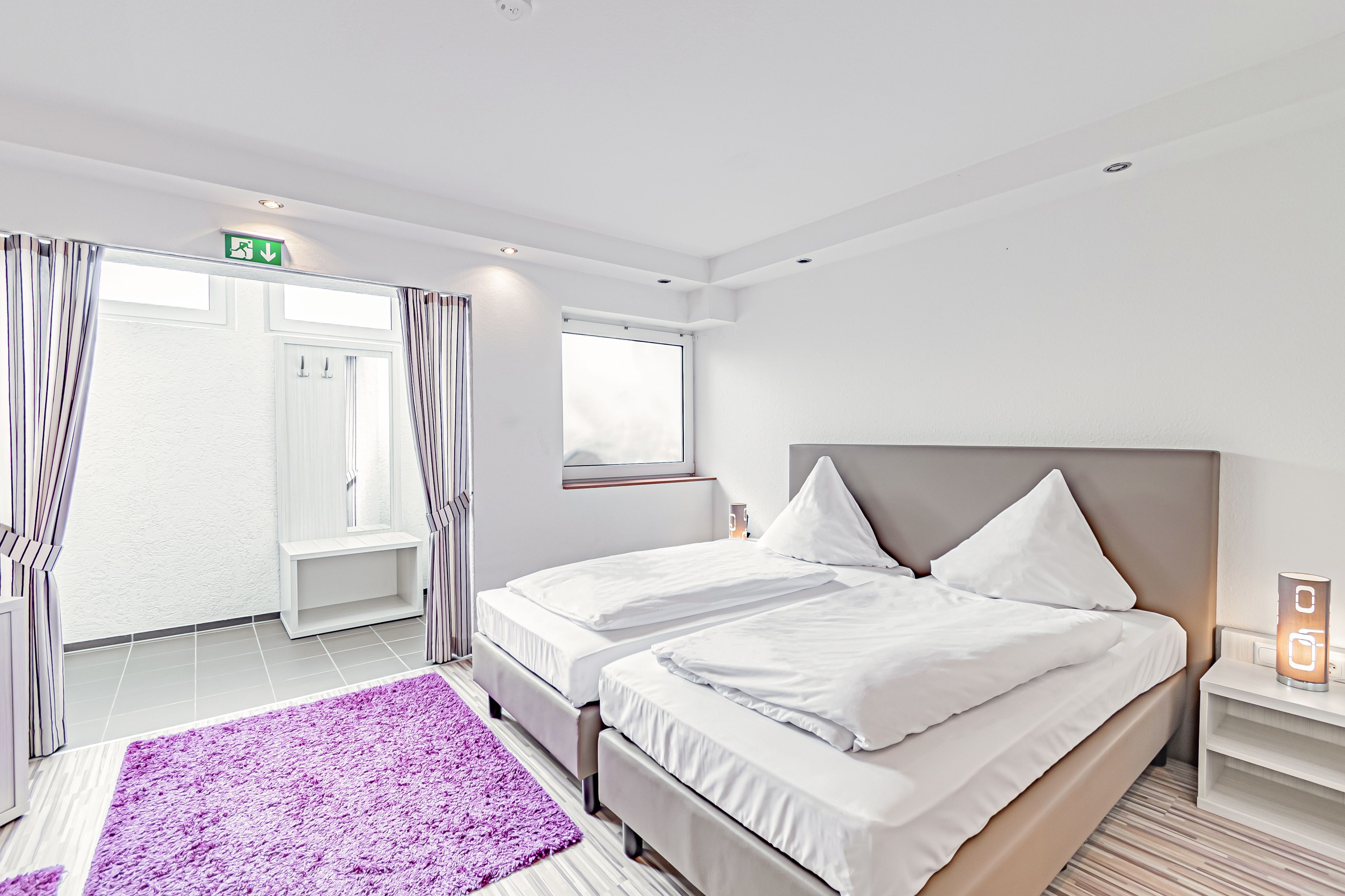 A modern hotel room with a large bed, a bedside table, a nightstand, a window, and a purple rug.
