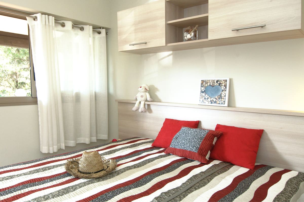 A bedroom with a striped bedspread, a hat on the bed, and a window with curtains.