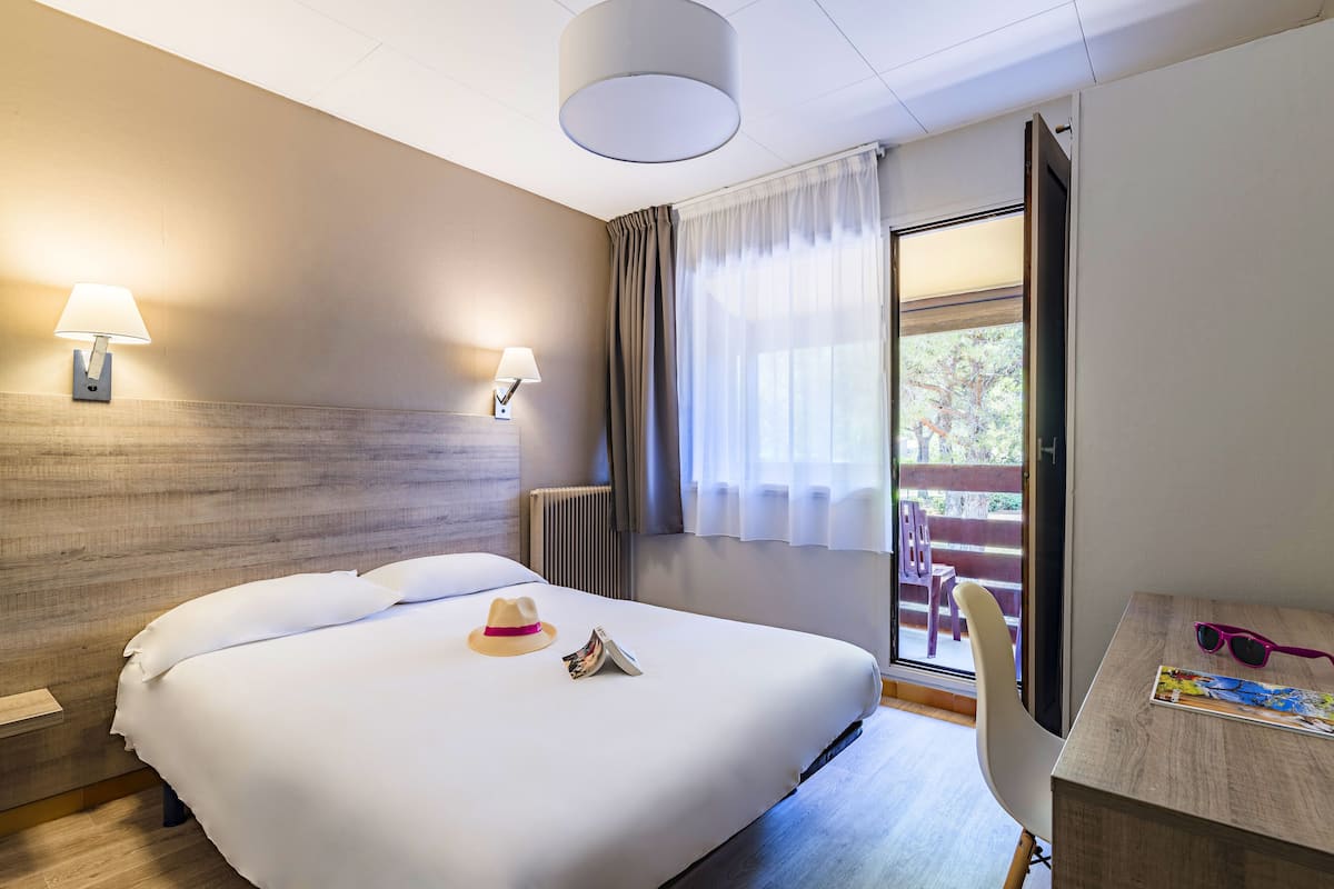 A hotel room with a large bed, two bedside lamps, a wooden headboard, a desk with a chair, and a view of the outdoors through a window with curtains.