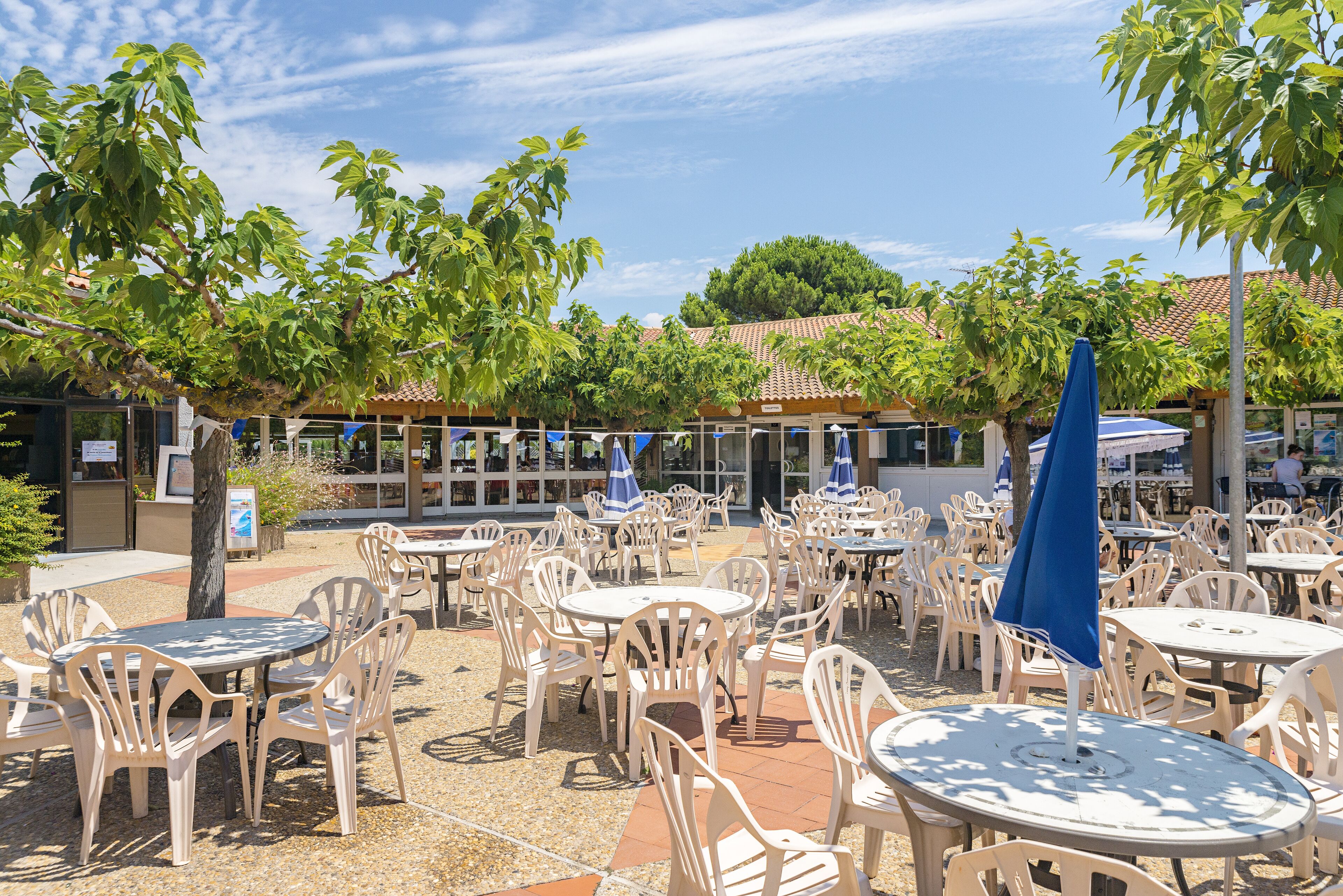 An outdoor café area with round tables and chairs, surrounded by trees and a building with a tiled roof in the background.