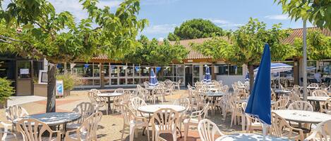 An outdoor café area with round tables and chairs, surrounded by trees and a building with a tiled roof in the background.