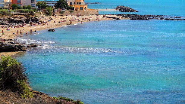 A coastal beach scene with buildings, clear blue water, and rocky outcrops.