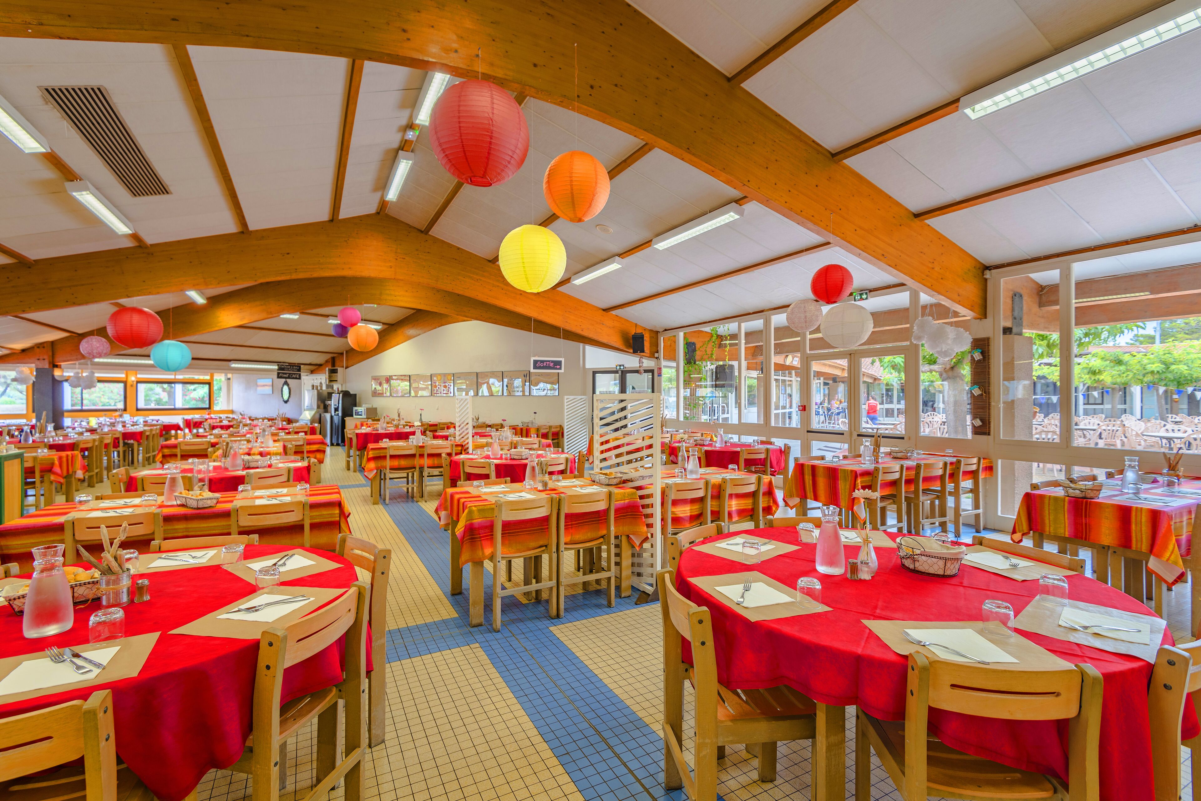 Un vaste espace de restauration intérieur, avec de nombreuses tables recouvertes de nappes rouges, des chaises en bois et des lanternes en papier suspendues.