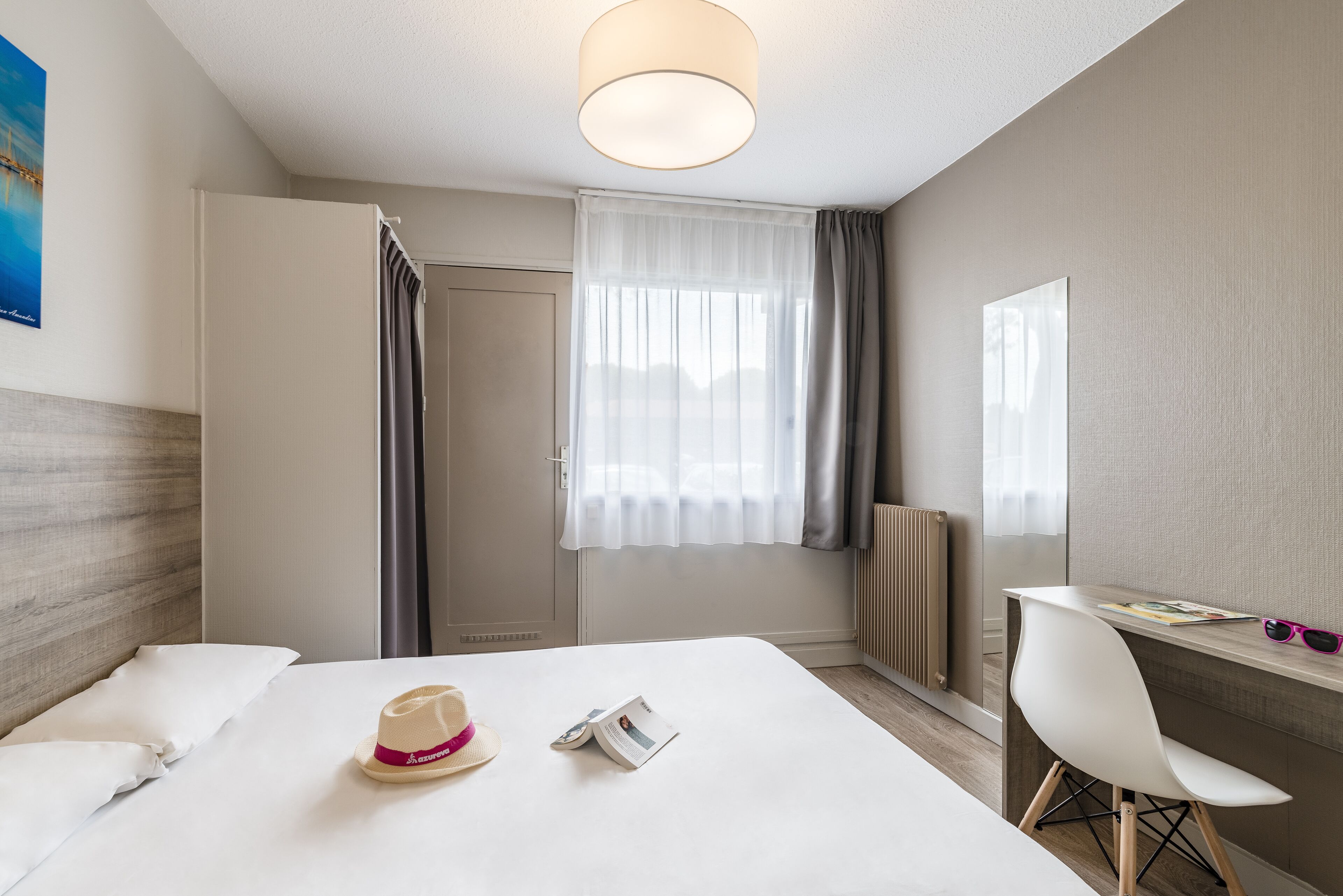 A hotel room with a bed, a desk, a chair, a window with curtains, and a radiator.