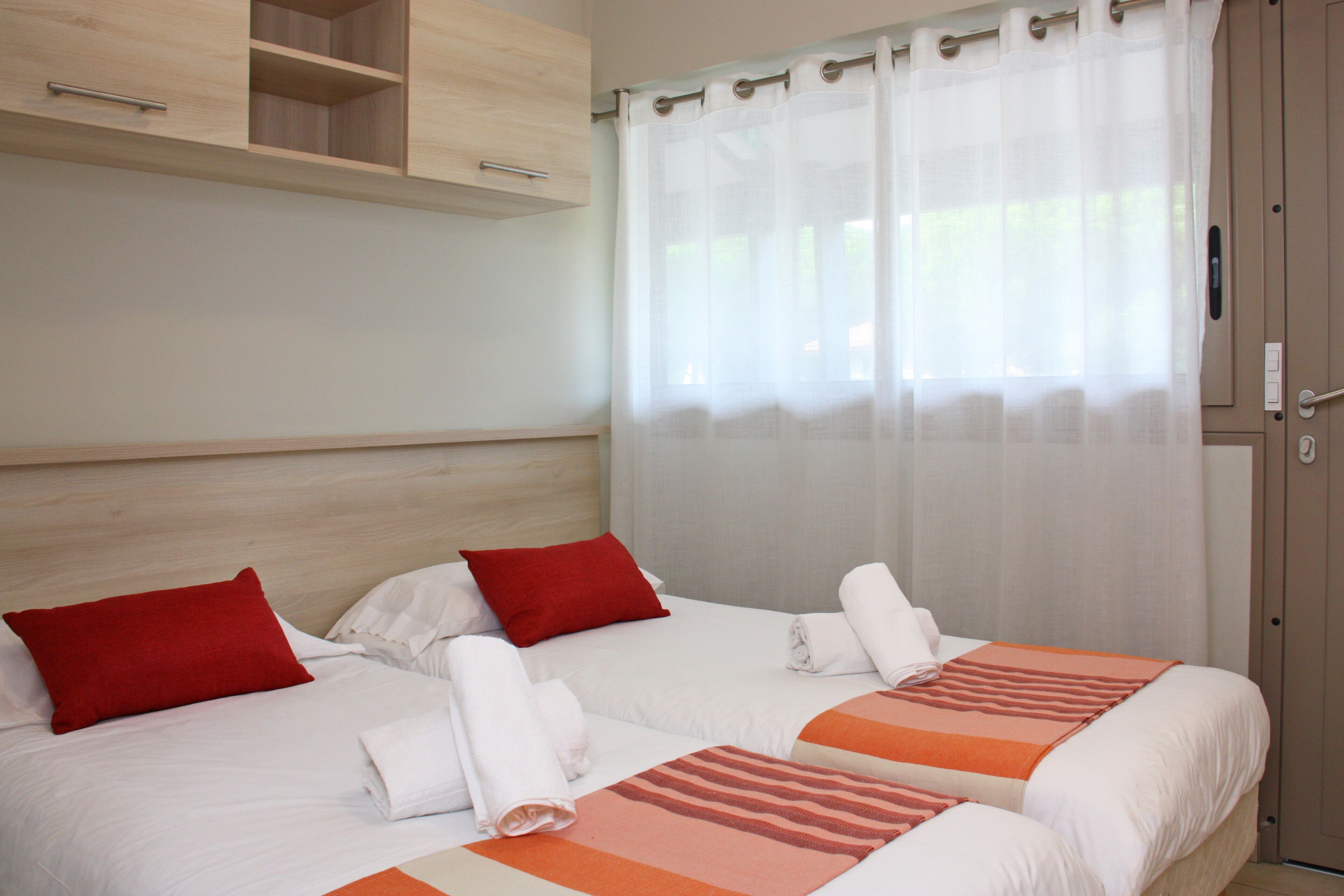 Une chambre d’hôtel avec deux lits, chacun agrémenté de draps blancs et d’oreillers rouges, d’une tête de lit en bois et d’une grande fenêtre ornée de rideaux légers.