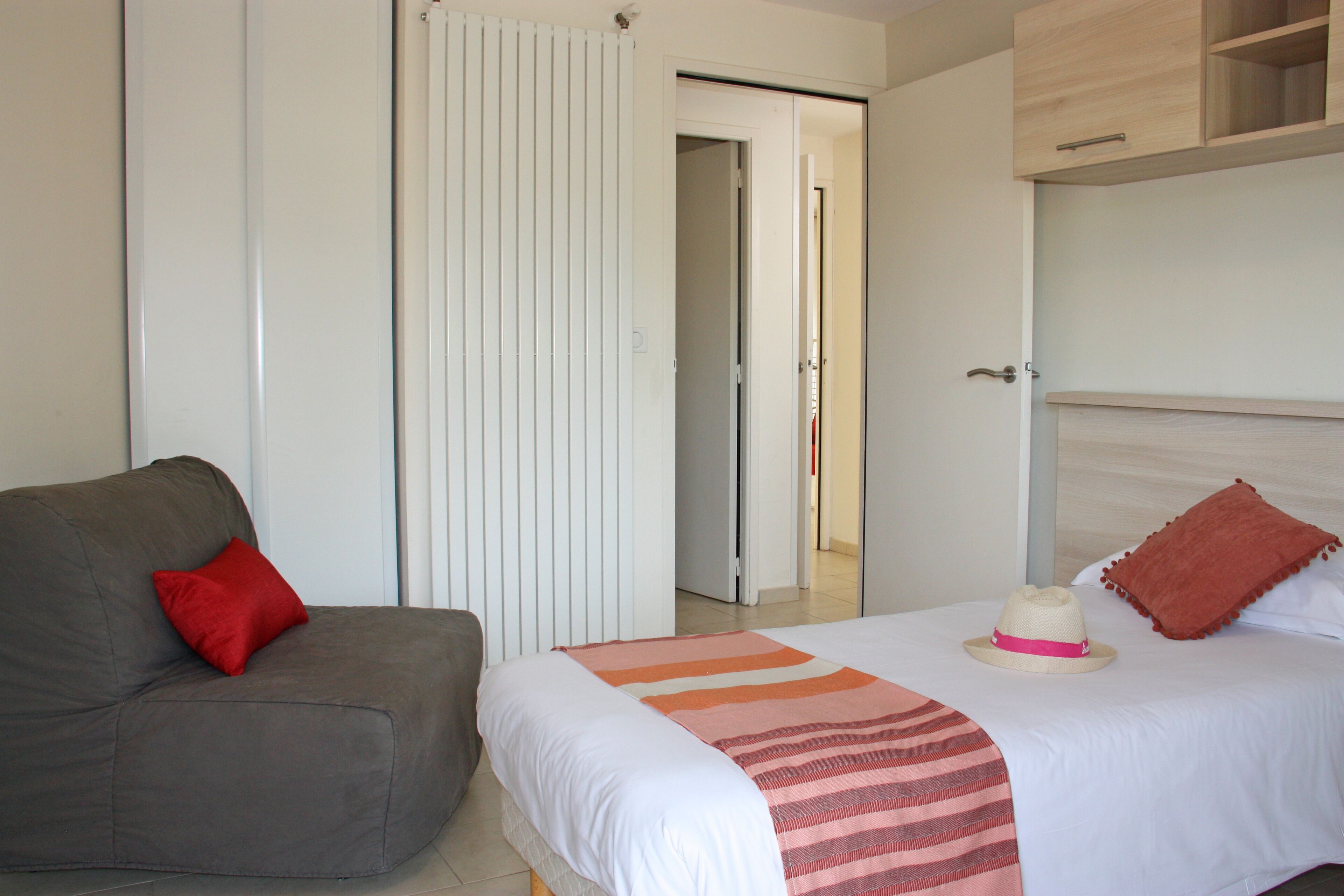 A bedroom with a bed, a grey sofa with a red pillow, a striped blanket, and a wooden wardrobe.