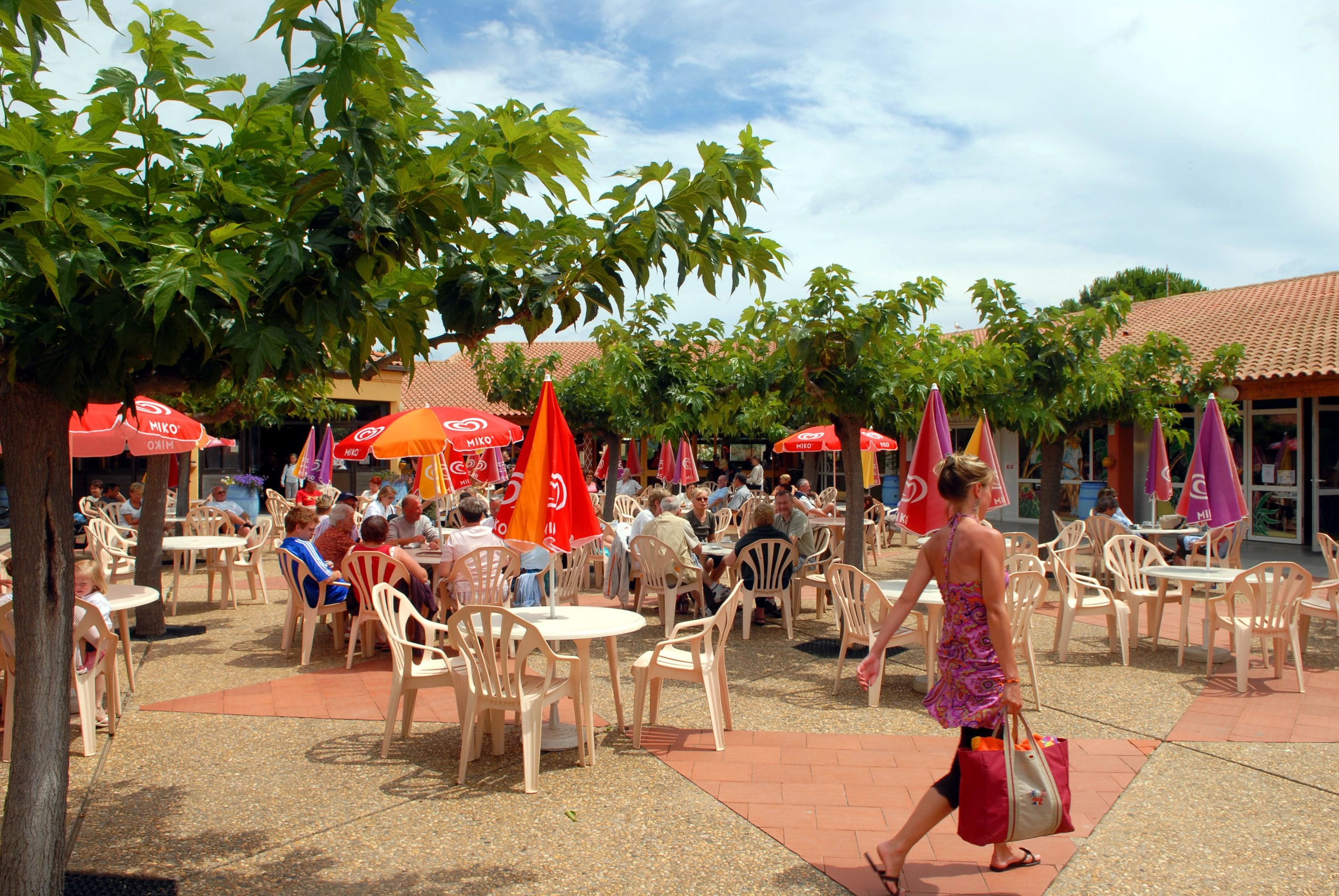 An outdoor cafe with numerous tables and chairs, surrounded by trees and umbrellas.