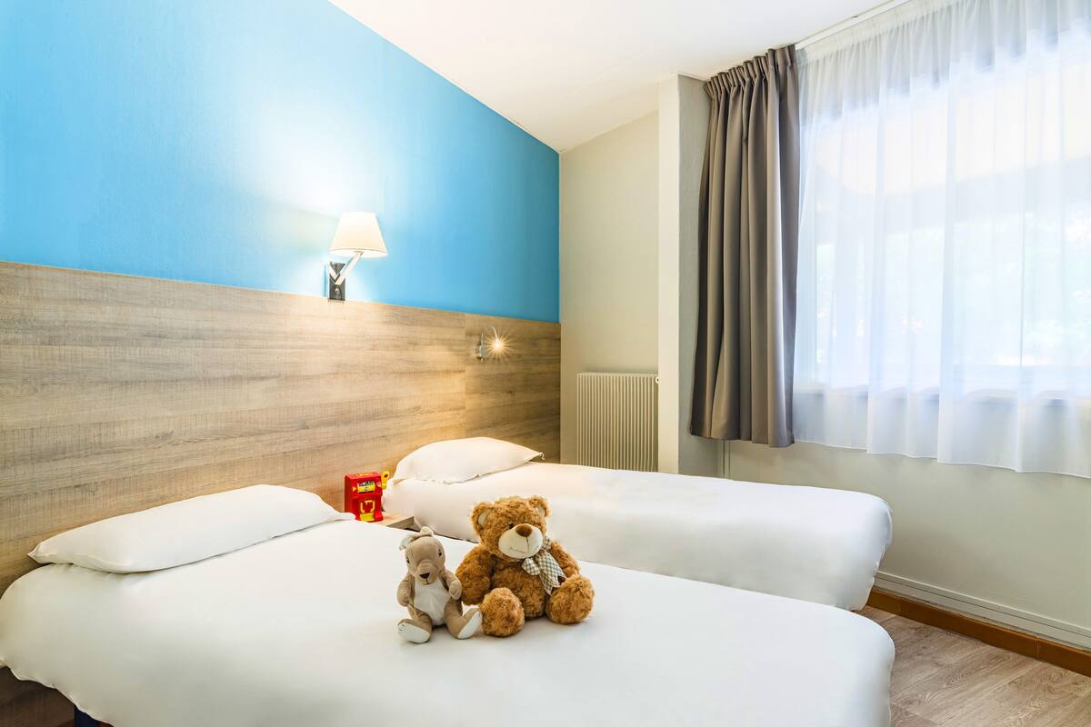 A hotel room with two beds, a wooden headboard, a nightstand with a lamp, and a window with curtains.