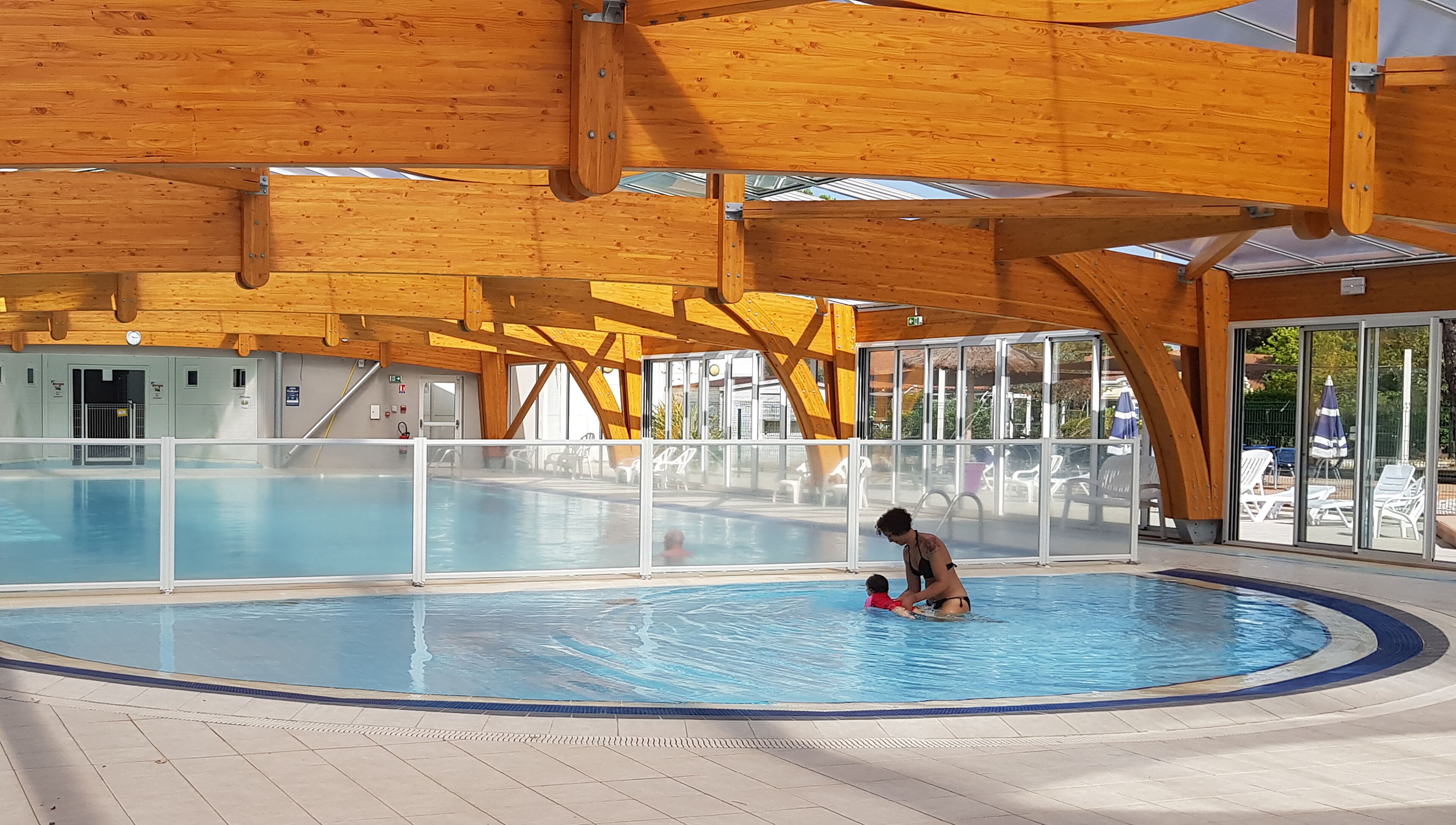 Une grande piscine intérieure dotée de poutres en bois, de murs en verre transparent et d’un sol carrelé.