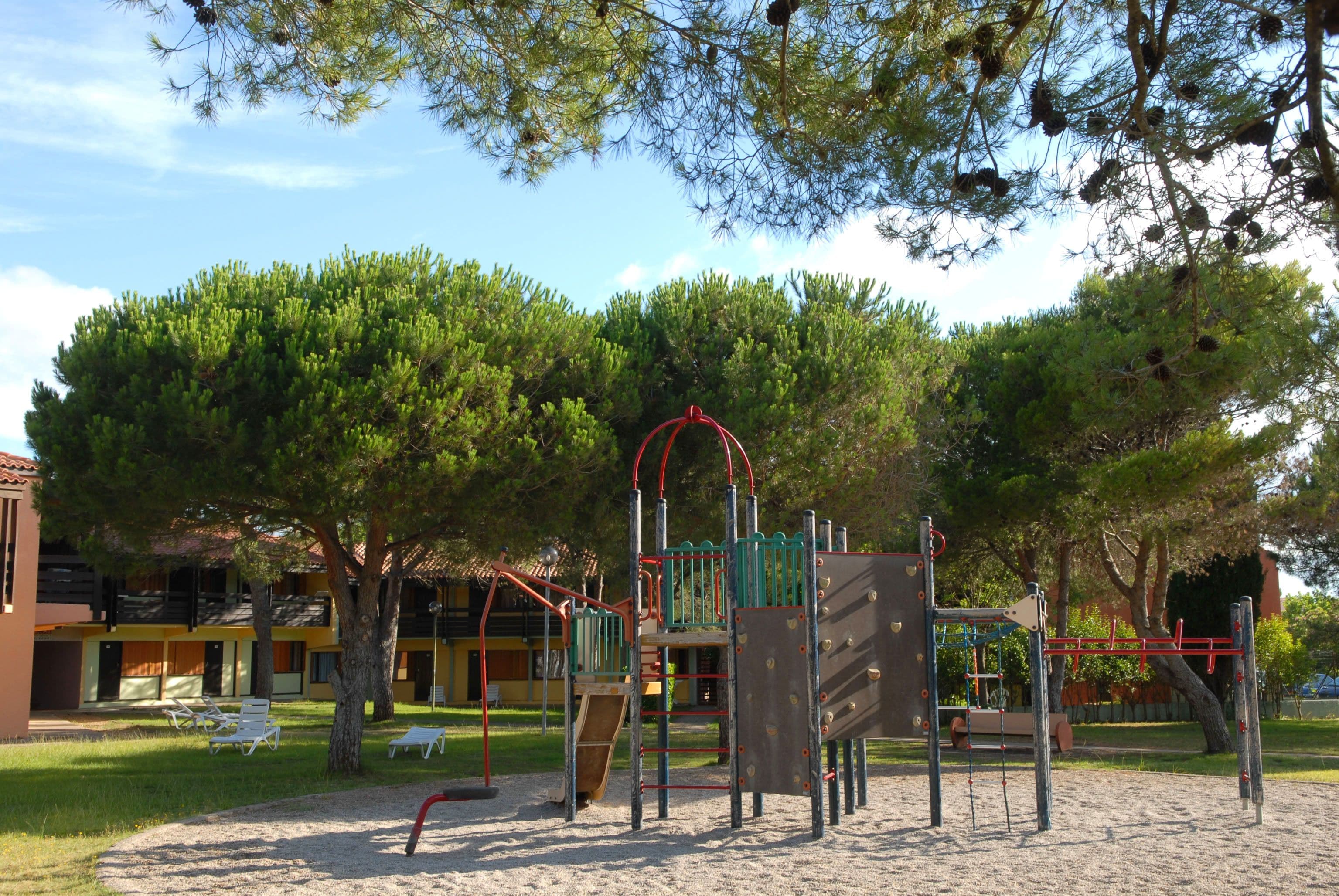 A playground with climbing structures and slides, surrounded by trees and a building with balconies.
