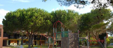 A playground with climbing structures and slides, surrounded by trees and a building with balconies.