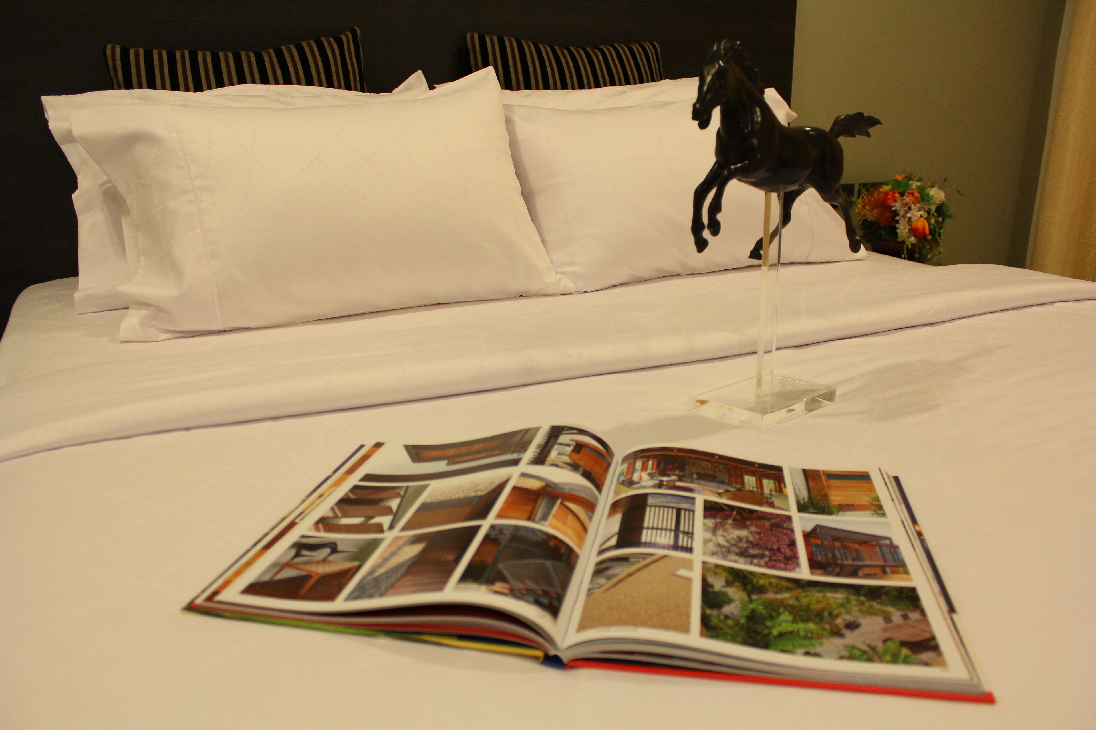 A neatly made bed with white linens, a decorative horse figurine, and a magazine with images of buildings.