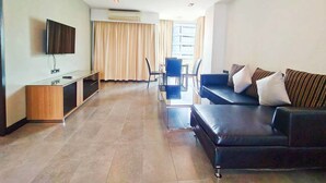 A modern living room with a flat-screen TV, a wooden entertainment unit, a black leather sofa, and a dining area with a glass table and chairs. - 14 Living (Bangkok)