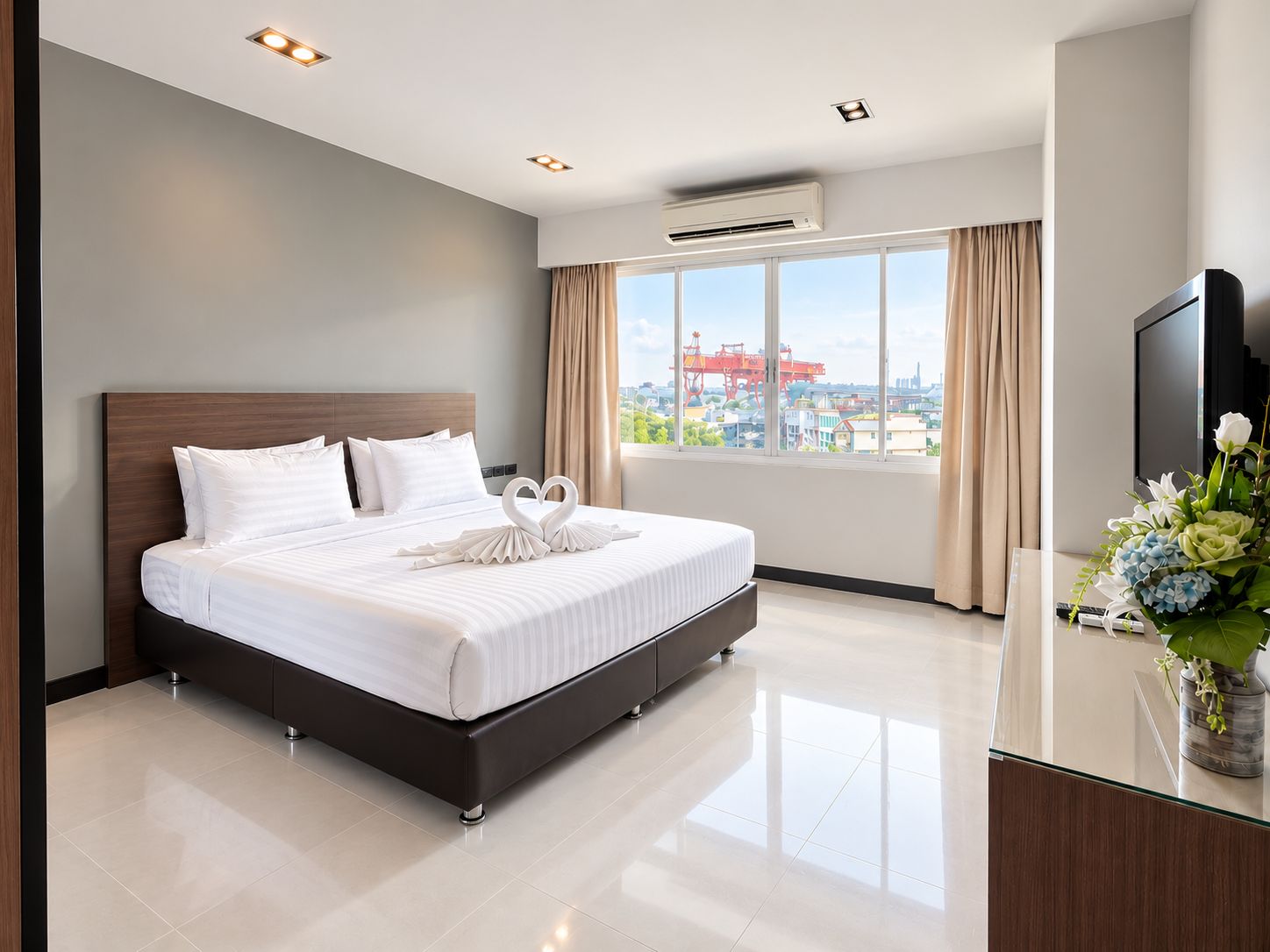 A hotel room with a large bed, a flat-screen TV, a desk with a vase of flowers, and a view of the cityscape through the window.
