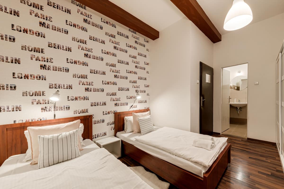 A hotel room with two beds, a wooden headboard, a wall with city names, a mirror, and a bathroom visible through an open door.