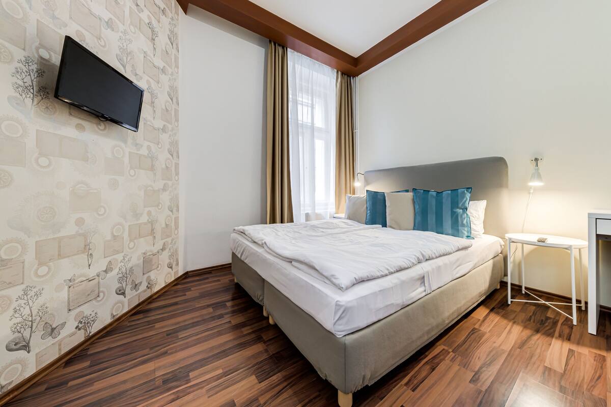 A bedroom with a bed, a TV mounted on the wall, a bedside table, and a window with curtains.