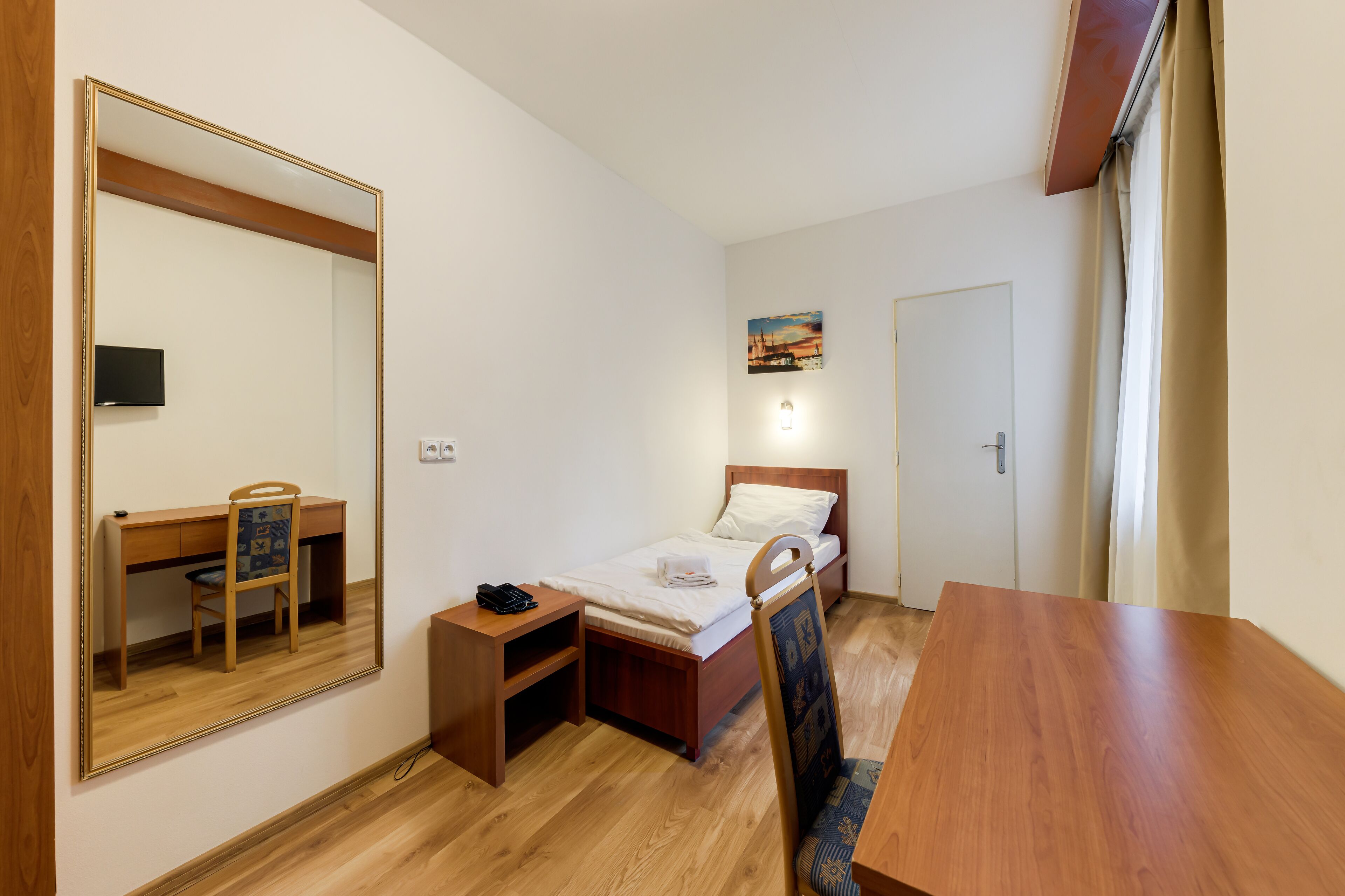 A small, tidy room with a wooden desk, a chair, a bed with a white mattress and pillows, a small bedside table with a phone, a wall-mounted TV, and a door with a light switch.