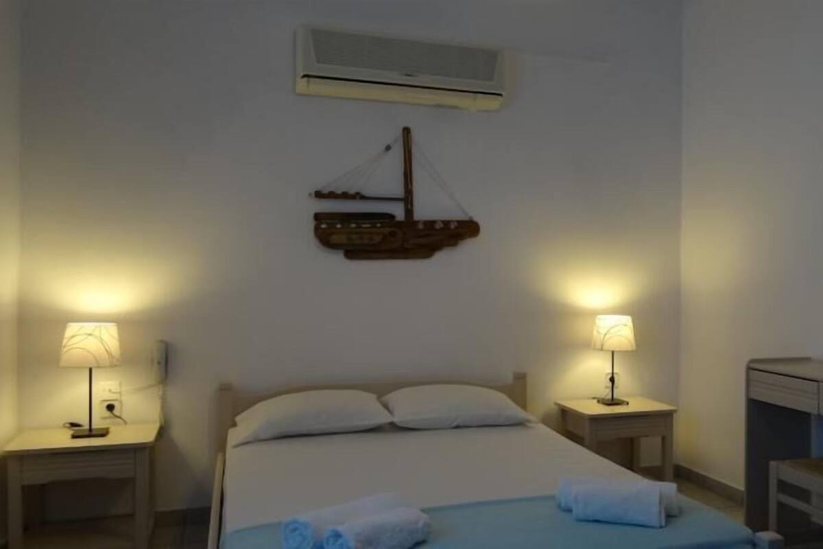 A hotel room with a bed, two bedside tables with lamps, a desk with a chair, an air conditioning unit, and a wall-mounted mirror.