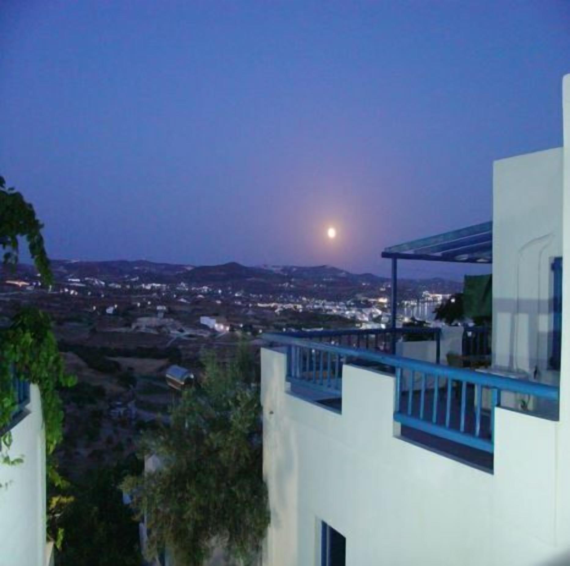 Vista nocturna de un pueblo costero con cielo iluminado por la luna, edificios modernos y un balcón con vistas al mar.