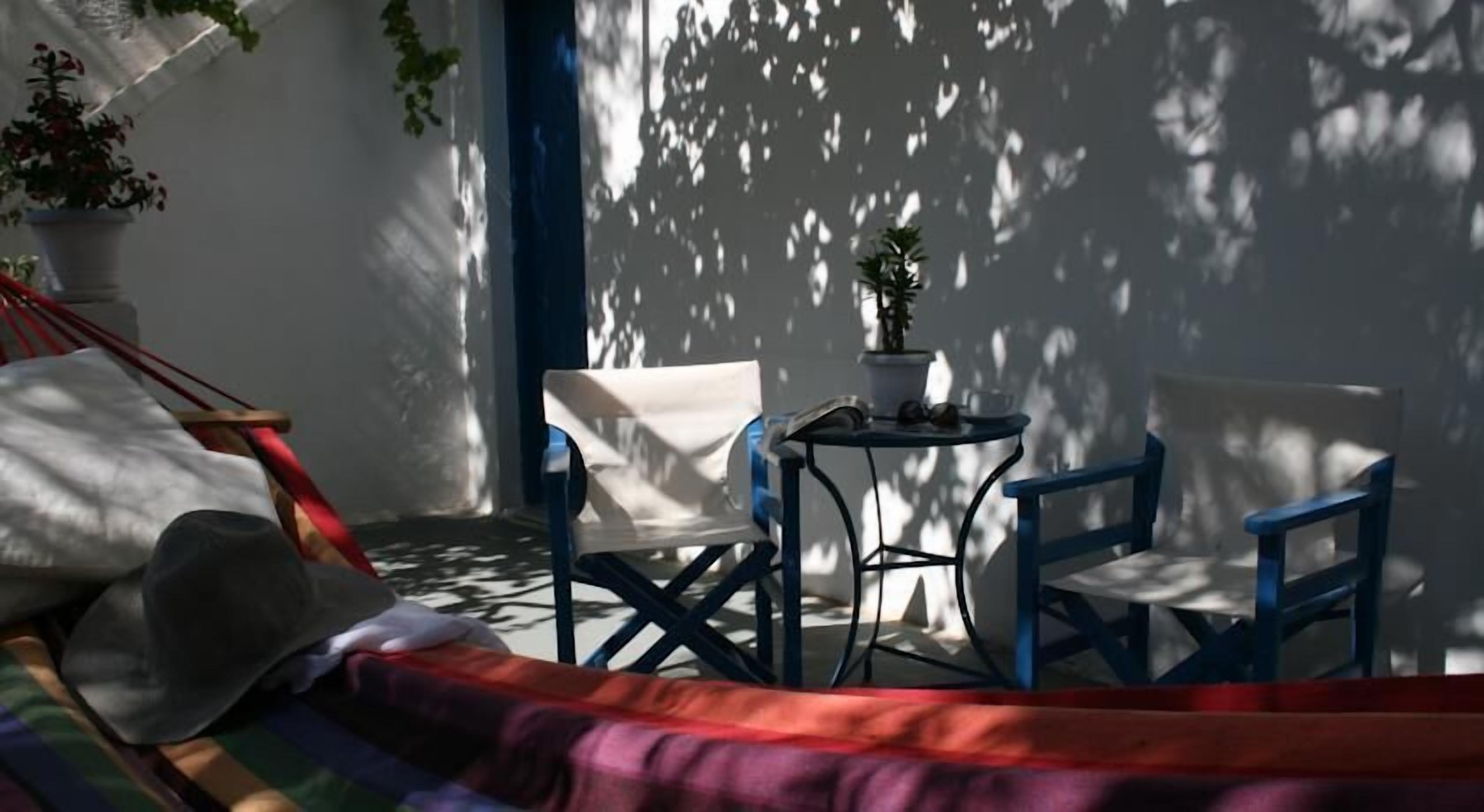 Una hamaca, dos sillas, una mesita con una planta y una pared blanca con sombras.