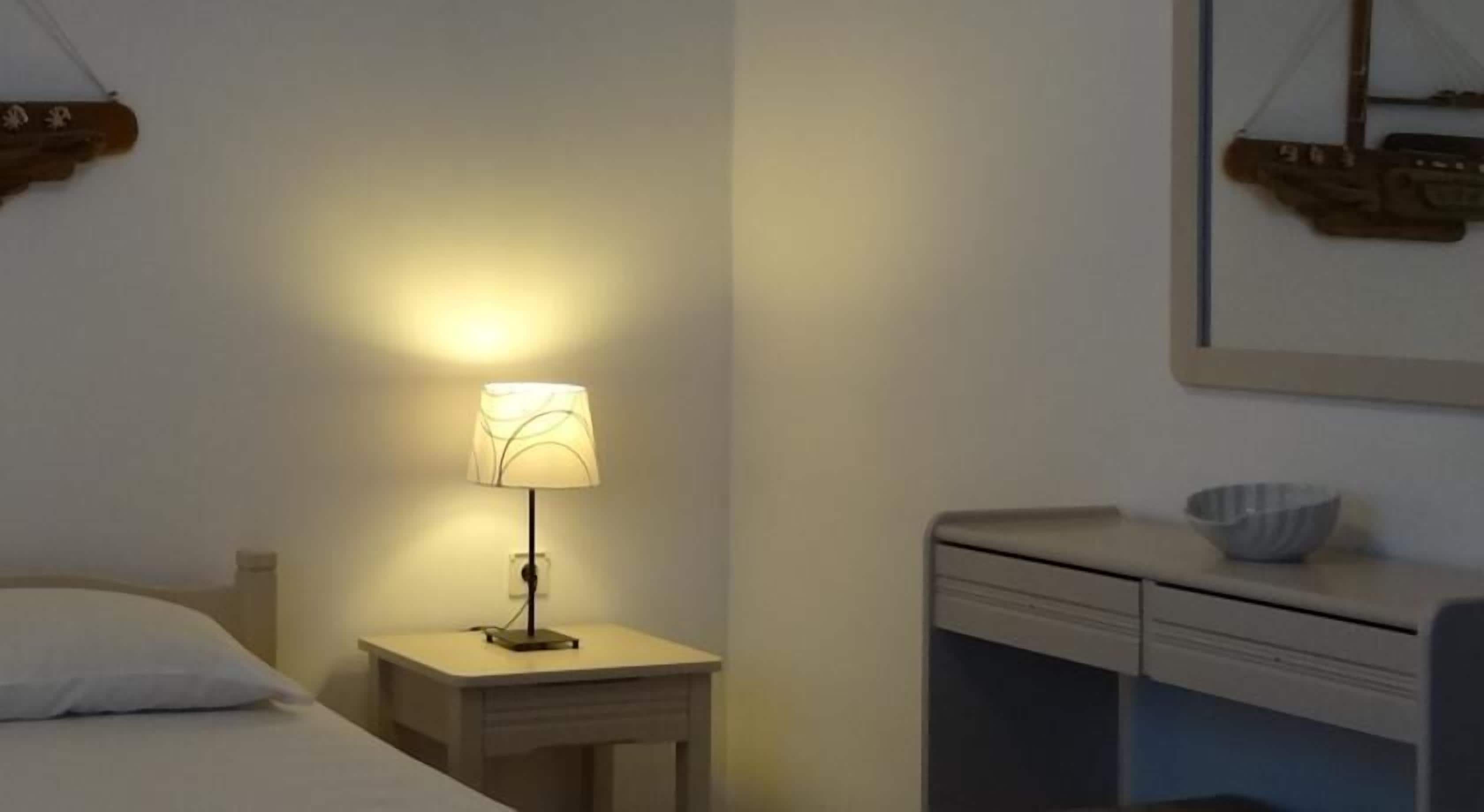 Habitación de hotel con cama, mesita de noche con lámpara, pequeño escritorio con un cuenco y estante de pared con objetos decorativos.