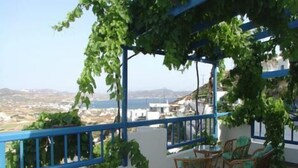 Un balcón con vistas al mar y las colinas, que cuenta con una pérgola cubierta de plantas trepadoras, una mesa con sillas y una barandilla azul.