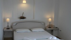 Una cama bien hecha con sábanas y almohadas blancas, flanqueada por dos mesitas de noche con lámparas, en una habitación sencilla y luminosa con paredes blancas y un pequeño adorno sobre la cama.