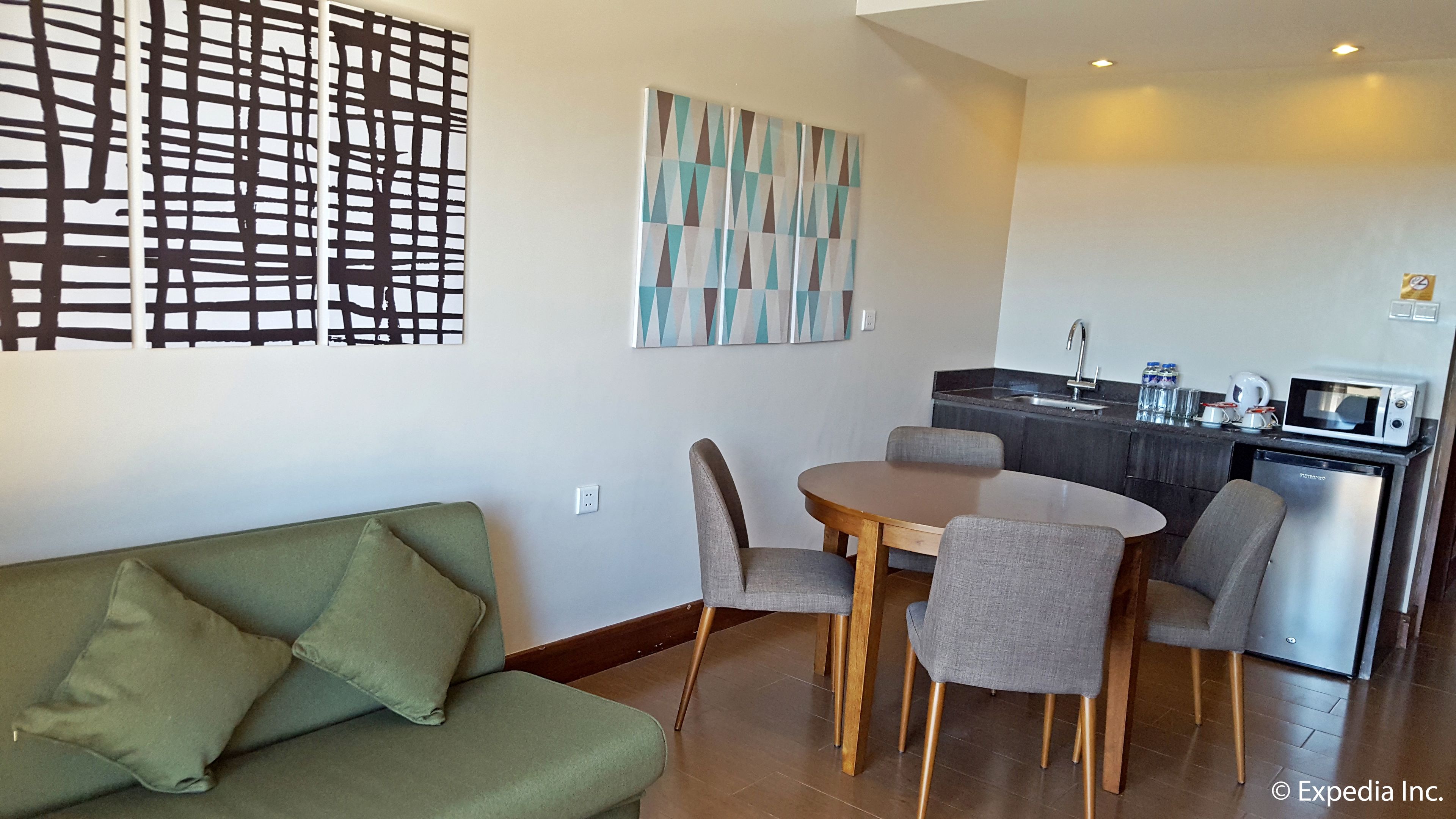 A modern living room with a green sofa, a wooden dining table with four chairs, and a kitchen area with a microwave and sink.