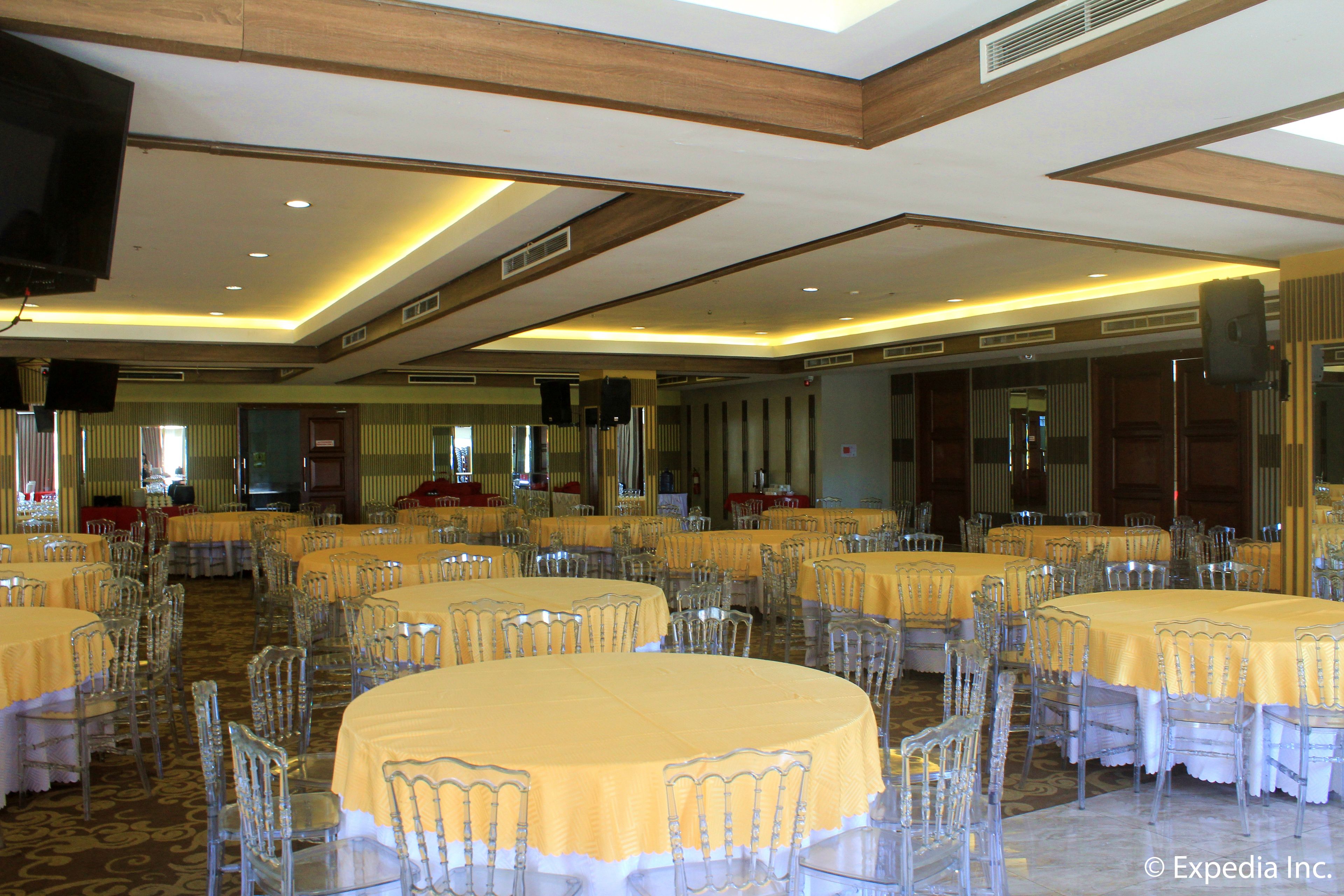 A banquet hall with round tables covered in yellow tablecloths and clear chairs, equipped with speakers and a television.