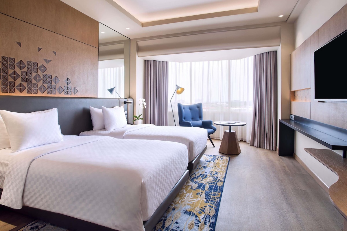 Quarto de hotel com duas camas, televisão, escrivaninha, cadeira e uma janela grande com cortinas.