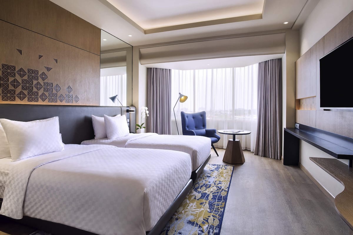 A hotel room with two beds, a TV, a desk, and a large window with curtains.