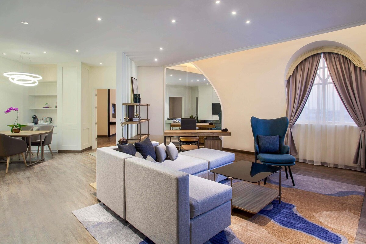 Una sala moderna con sofá, sillón y mesa de centro, un comedor con mesa y sillas, y una cocina visible a través de un arco.