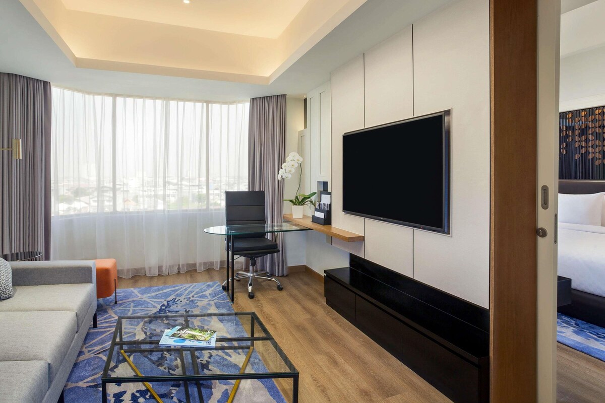 A hotel room with a sofa, a glass coffee table, a TV, a desk with a chair, and a bed.