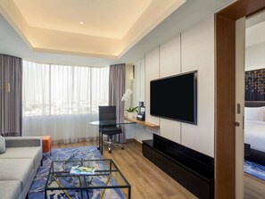 A hotel room with a sofa, a glass coffee table, a TV, a desk with a chair, and a bed. - Mercure Jakarta Batavia (Jakarta)