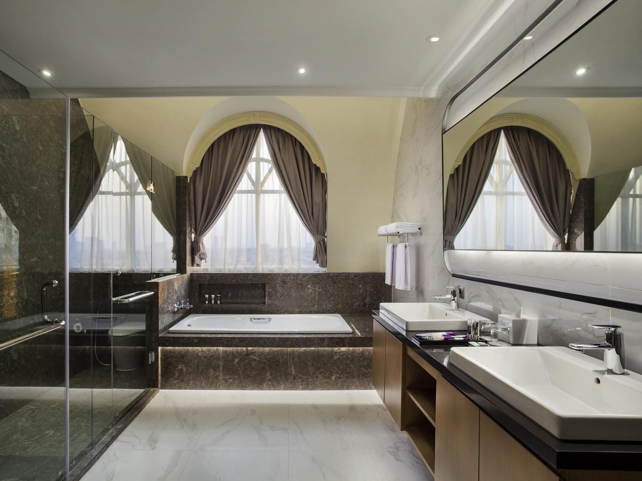 A modern bathroom with a bathtub, two sinks, a glass shower enclosure, and large windows with curtains.